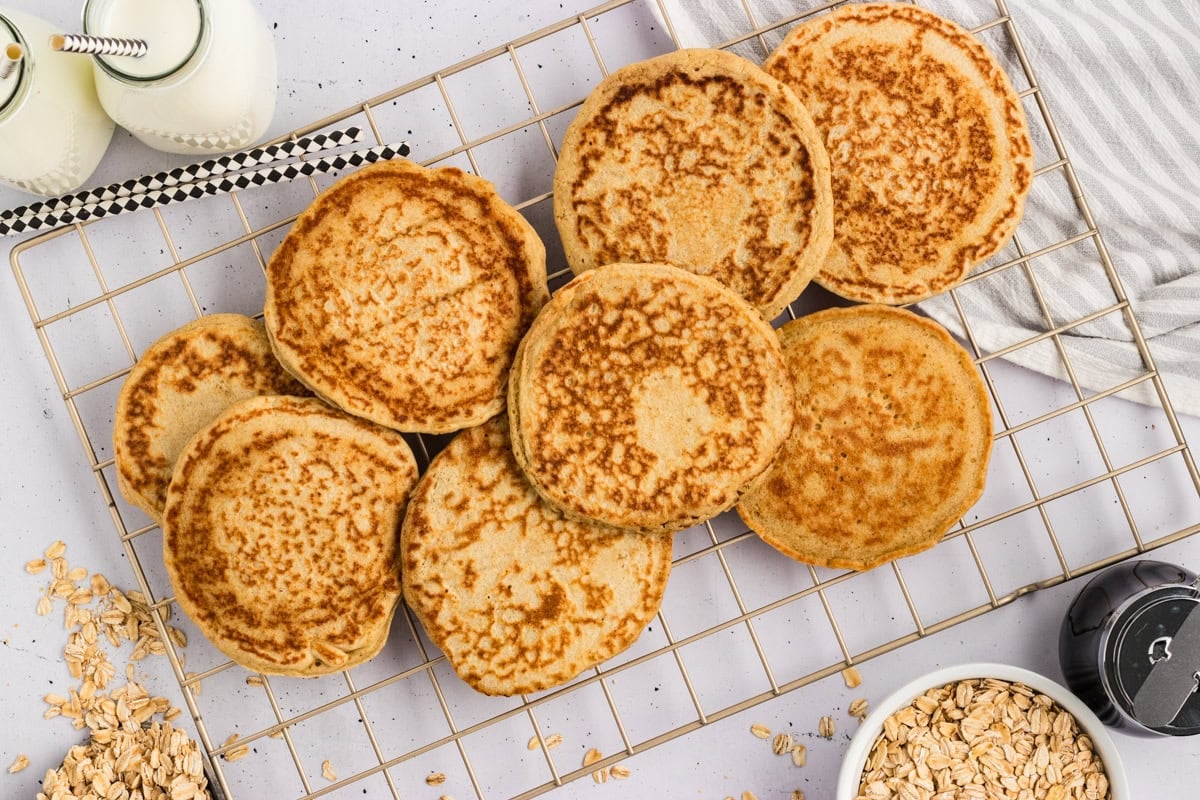Cooked oat flour pancakes cooling on a wire rack next to milk bottles and a bowl of rolled oats.
