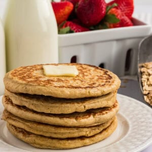 Stack of oat flour pancakes topped with a pat of butter, served with fresh strawberries and a bottle of milk in the background.