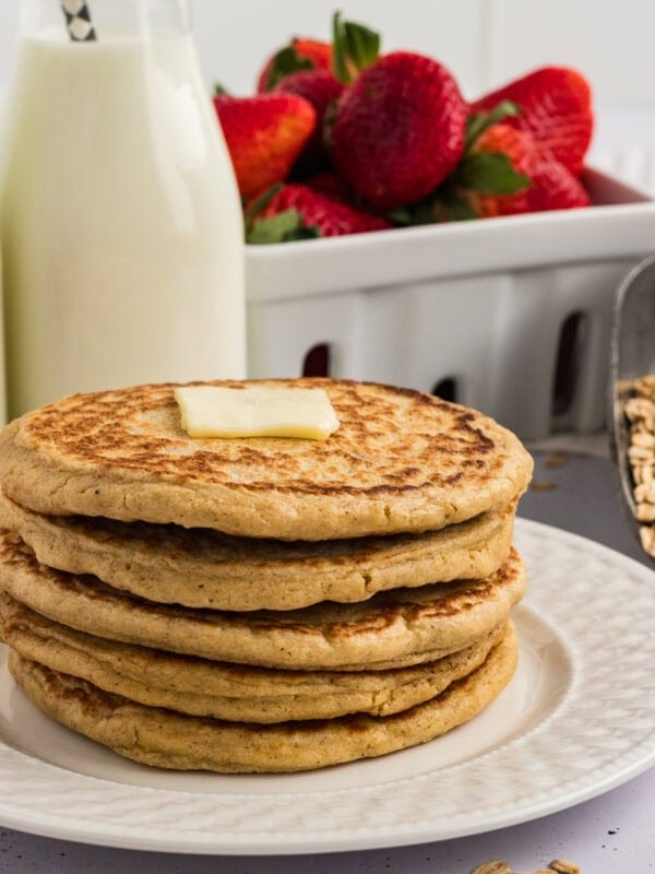 Stack of oat flour pancakes topped with a pat of butter, served with fresh strawberries and a bottle of milk in the background.