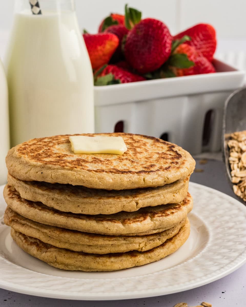 Stack of oat flour pancakes topped with a pat of butter, served with fresh strawberries and a bottle of milk in the background.