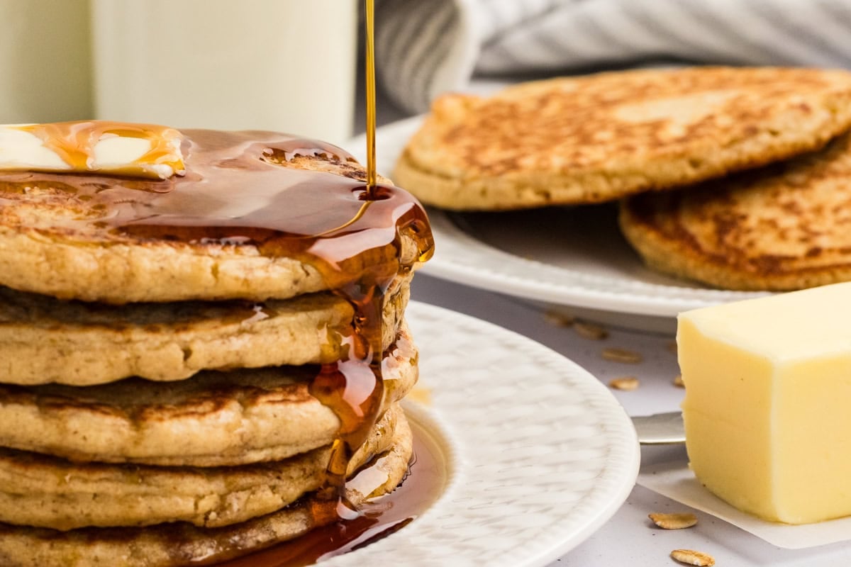 Maple syrup pouring over a stack of buttered oat flour pancakes.