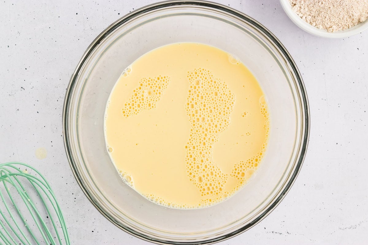 Milk, eggs, and vanilla whisked together in a glass bowl for oat flour pancake batter.