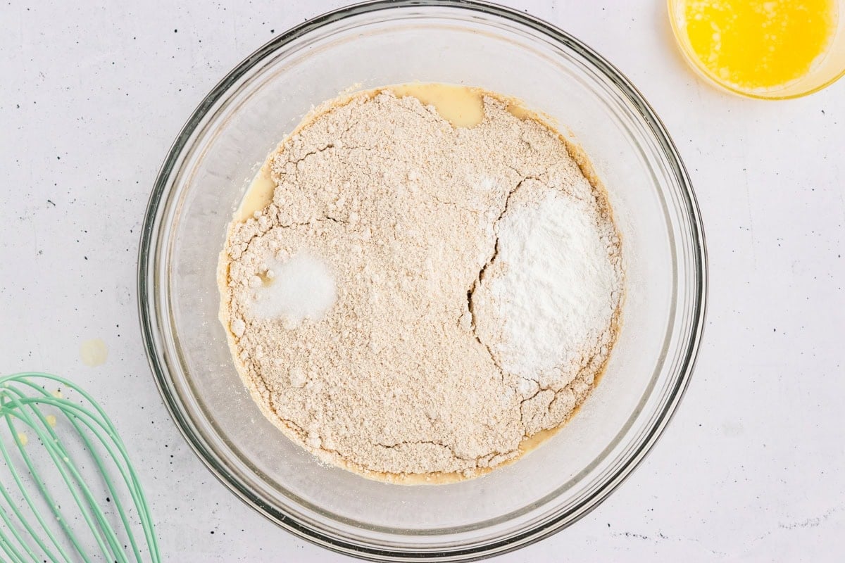 Oat flour, baking powder, and salt added on top of the whisked wet ingredients in a glass mixing bowl.