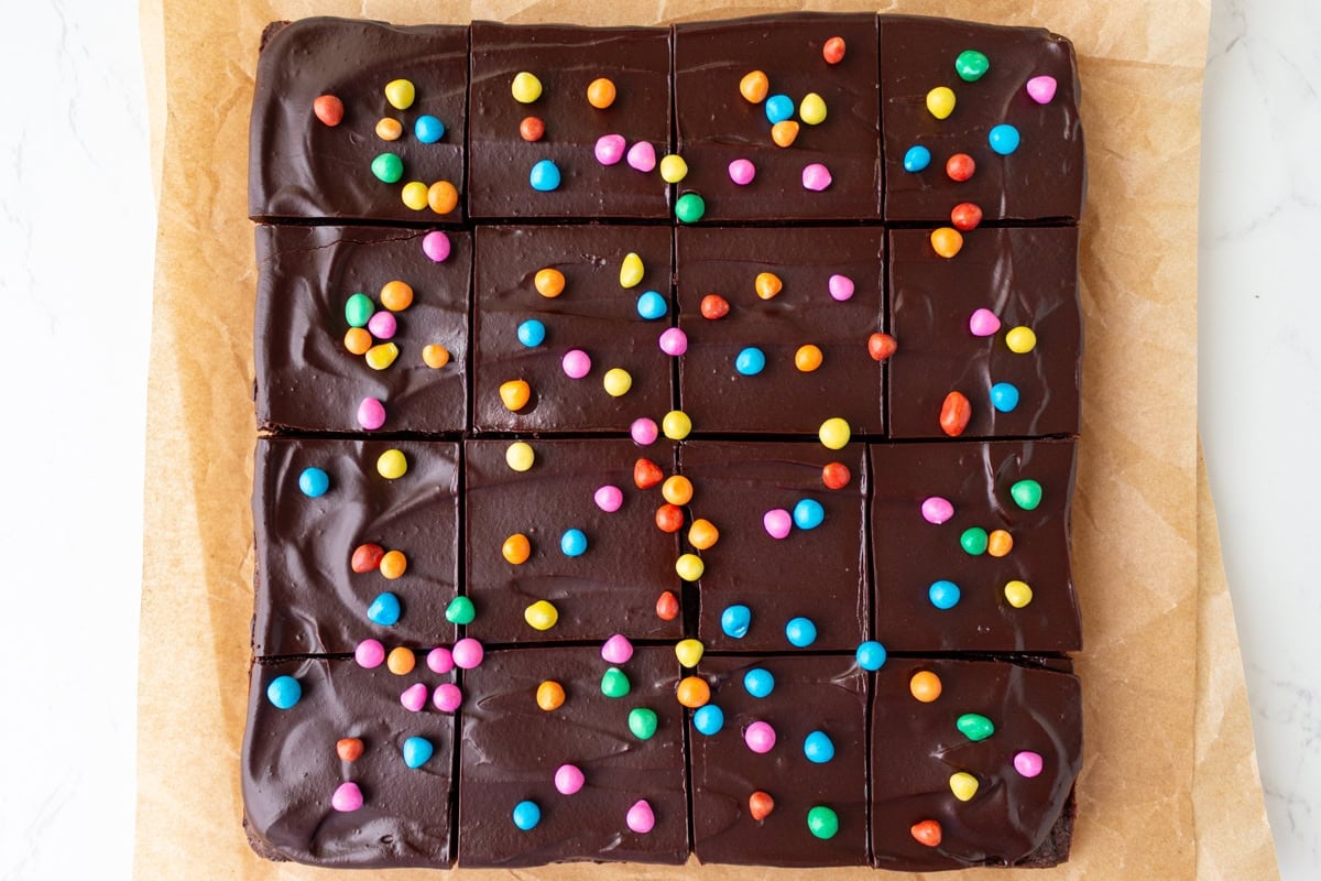 Frosted gluten-free cosmic brownies cut into squares and topped with rainbow candy chips on parchment paper.