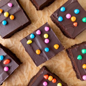 Overhead shot of gluten-free cosmic brownie squares topped with chocolate ganache and rainbow candy chips scattered on parchment paper.