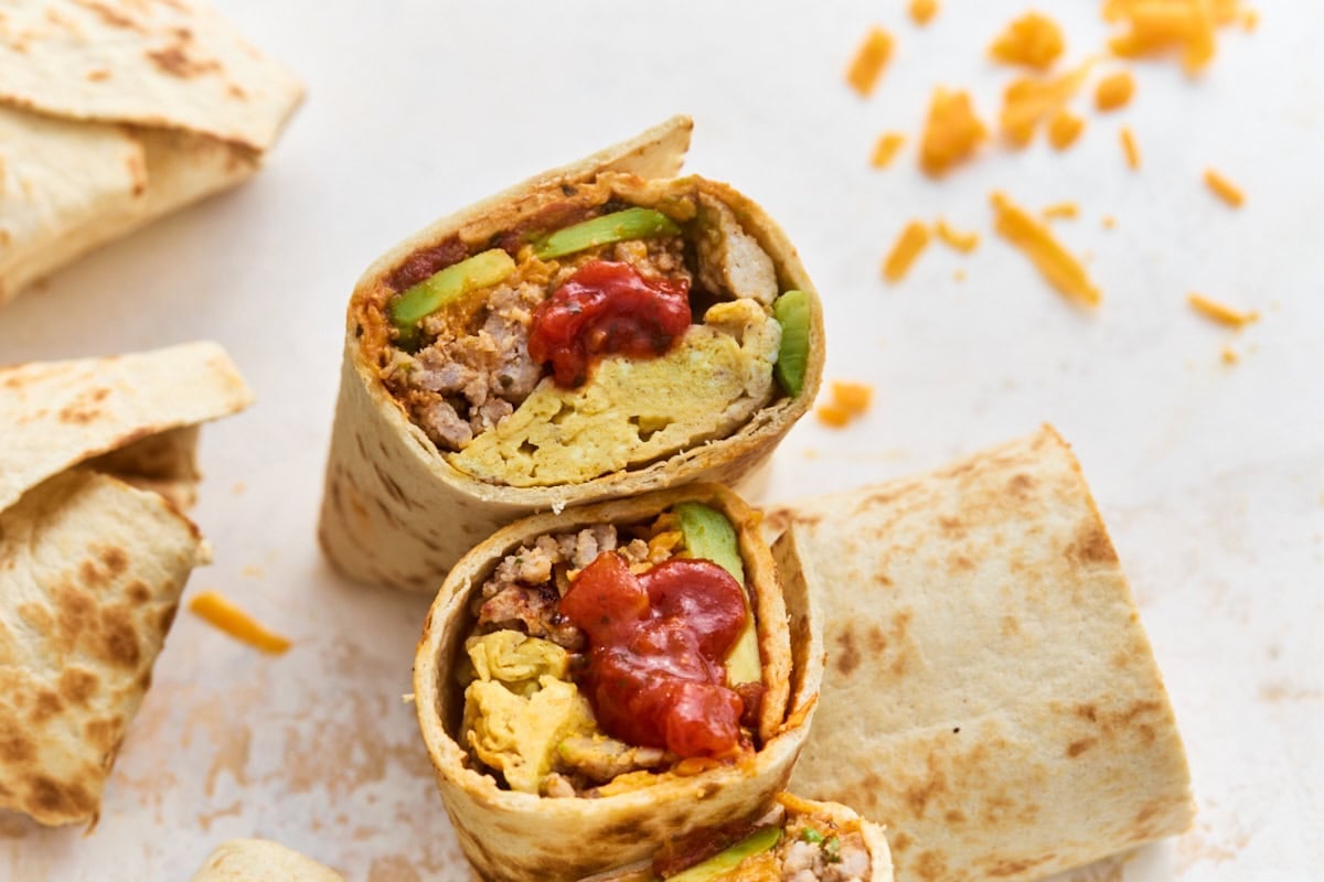 Close-up of a gluten-free breakfast burrito sliced in half, showing layers of eggs, sausage, cheese, avocado, and salsa
