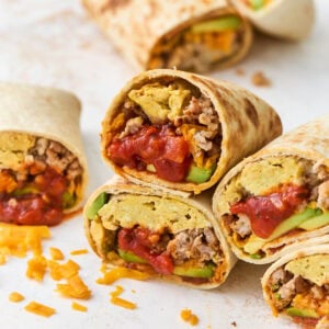Gluten-free breakfast burritos cut in half and stacked, showing scrambled eggs, sausage, melted cheddar, avocado, and salsa inside a golden tortilla