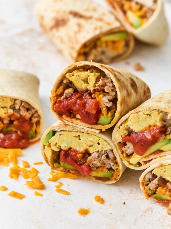 Gluten-free breakfast burritos cut in half and stacked, showing scrambled eggs, sausage, melted cheddar, avocado, and salsa inside a golden tortilla