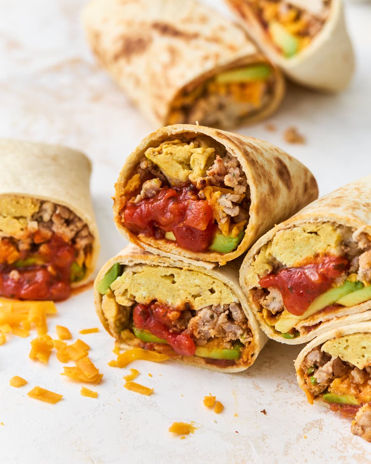 Gluten-free breakfast burritos cut in half and stacked, showing scrambled eggs, sausage, melted cheddar, avocado, and salsa inside a golden tortilla