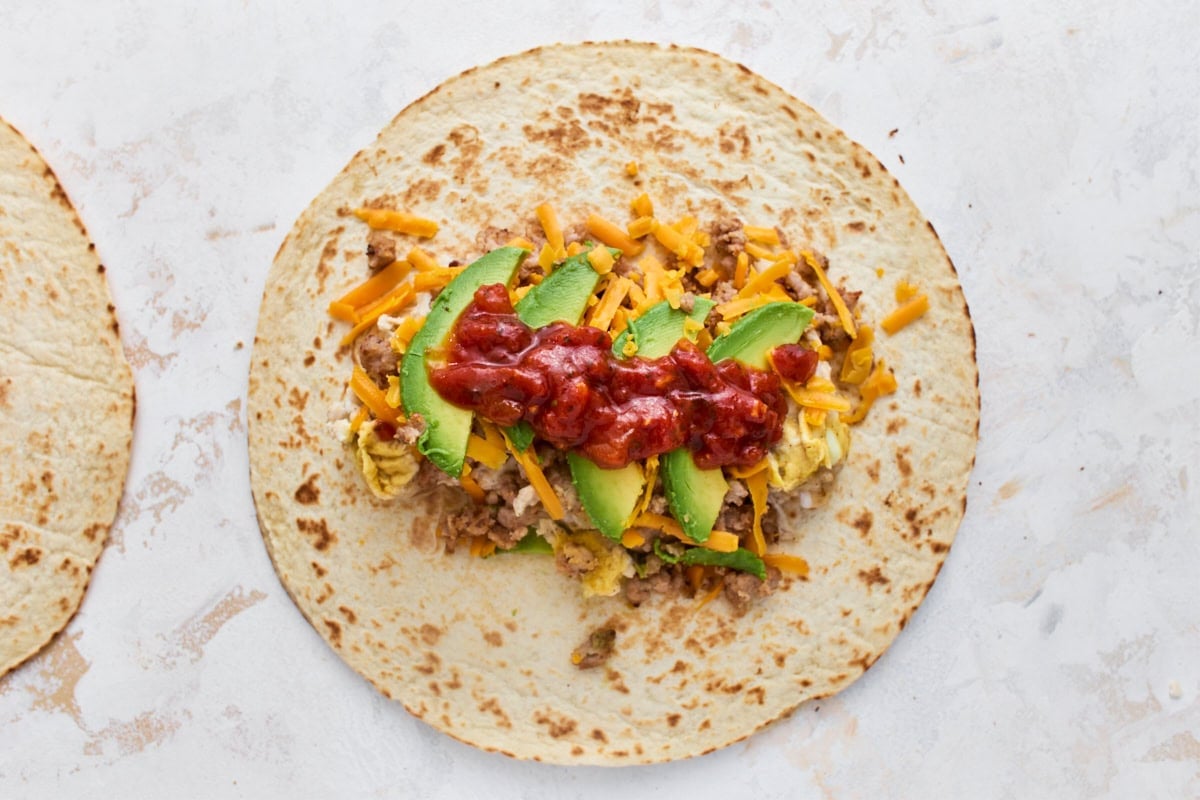 Gluten-free tortilla topped with scrambled eggs, sausage, shredded cheddar, avocado slices, and salsa before rolling