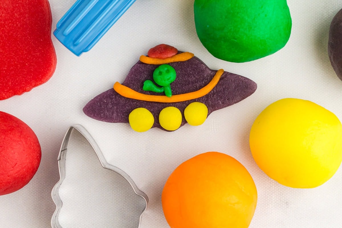 a purple UFO with a green alien, orange trim, and yellow circles, made from gluten free play doh