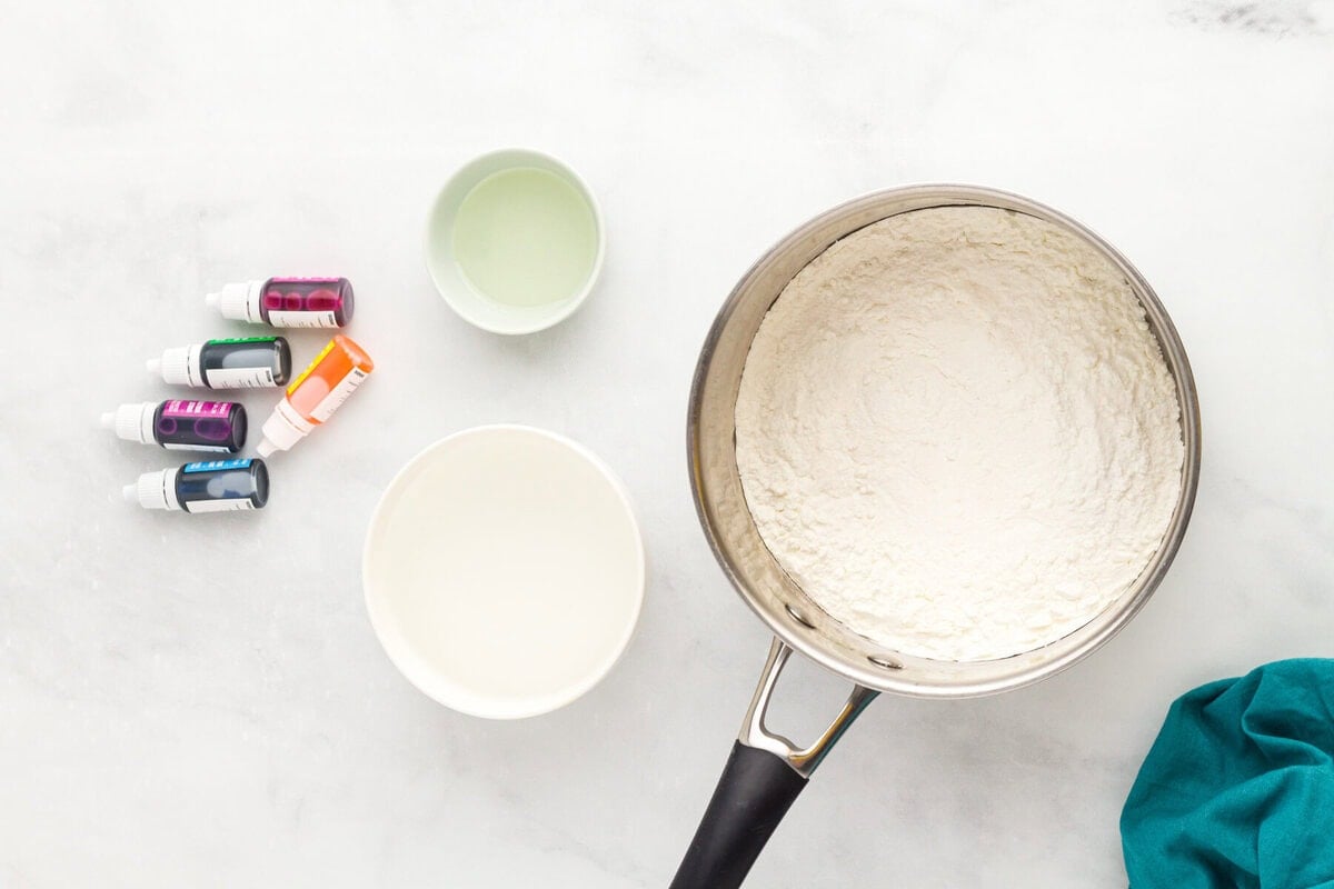 flour mixture in a pot with bowls of water and oil, and food coloring bottles nearby
