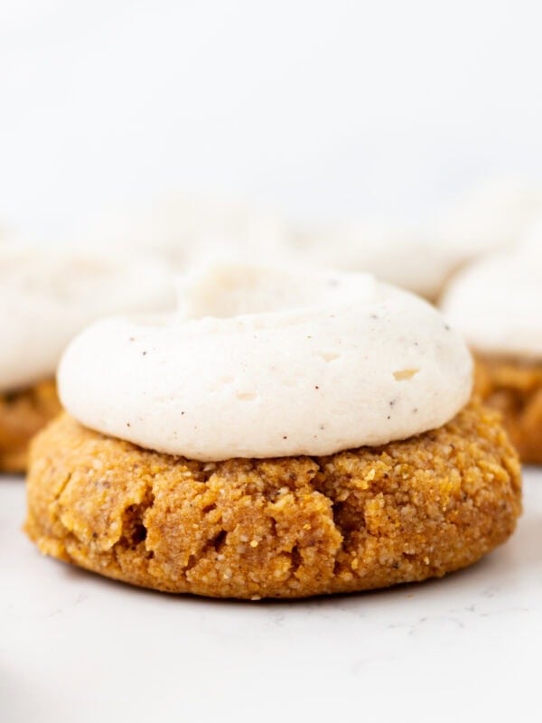 Gluten-free almond flour pumpkin cookie topped with brown butter cream cheese frosting, with more frosted cookies blurred in the background.