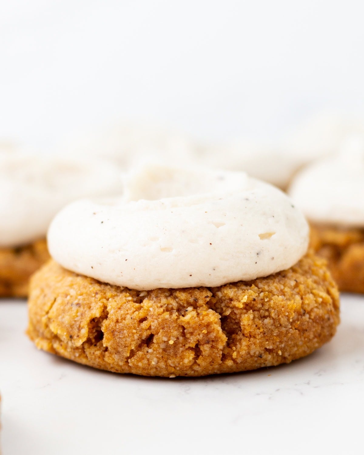 Gluten-free almond flour pumpkin cookie topped with brown butter cream cheese frosting, with more frosted cookies blurred in the background.